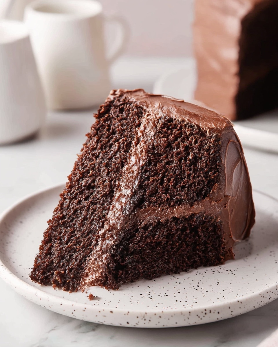 A close-up of a thick slice of two-layer chocolate cake on a white plate with small black speckles, resting on a white marbled surface. Each dark brown cake layer looks moist and spongy, separated and covered by a shiny, smooth layer of rich chocolate frosting that also coats the top and sides of the slice. The frosting has a slightly wavy texture with soft peaks and ridges, adding a creamy look. In the blurred background, there are white ceramic objects that hint at a cozy setting. Photo taken with an iphone --ar 4:5 --v 7
