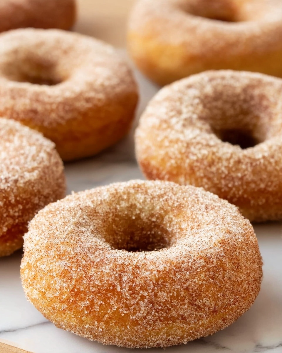 The image shows several round donuts with a golden-brown color, each covered evenly in a layer of sugar and cinnamon granules giving a coarse, grainy texture. The donuts have a visible hole in the middle and appear soft and fluffy with a slightly rough surface due to the sugar coating. They are placed closely on a white marbled surface, with the focus on one donut in the front while others are slightly blurred in the background. photo taken with an iphone --ar 4:5 --v 7