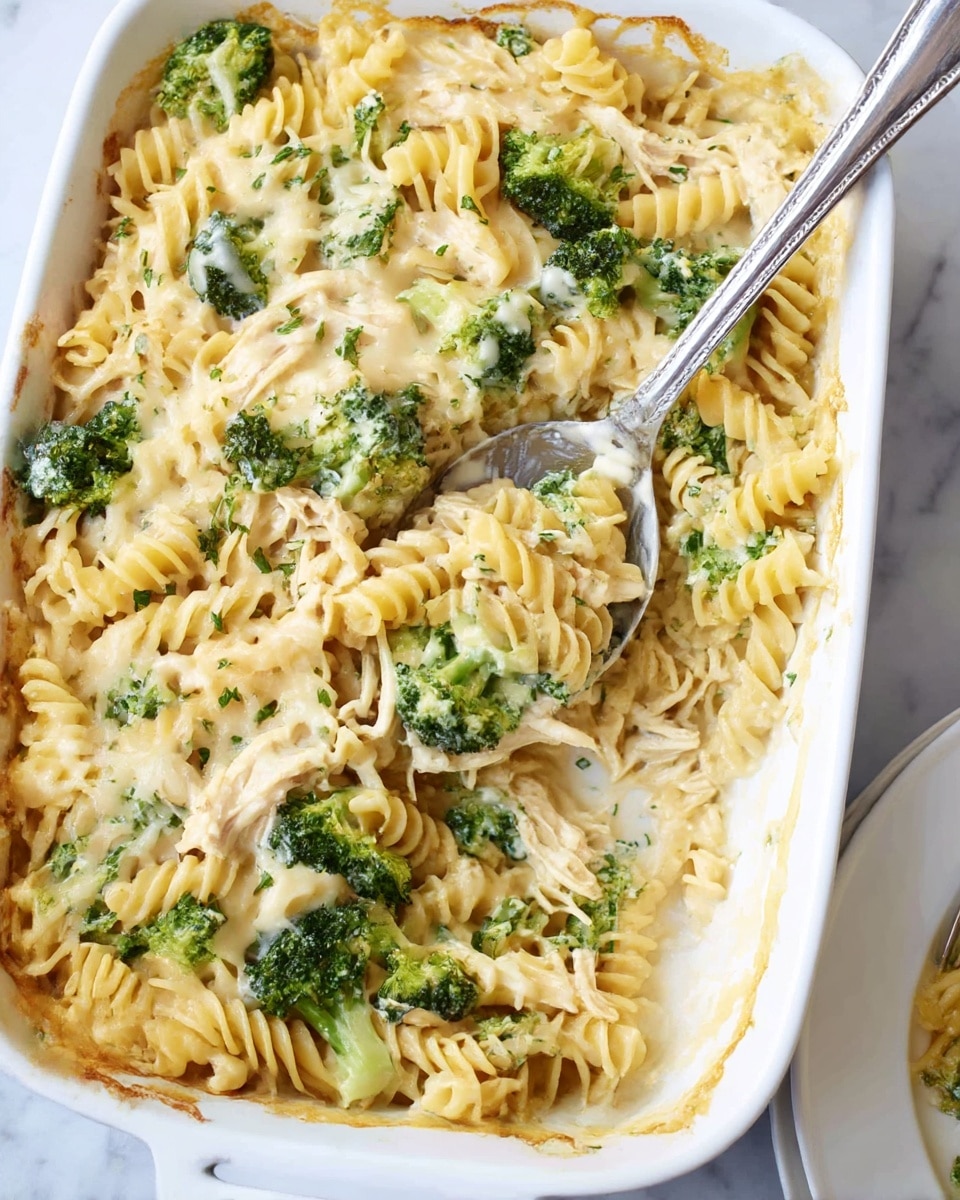 A white baking dish filled with a creamy pasta casserole, showing at least three layers: a base of yellow spiral pasta, mixed with green broccoli pieces, topped with a light cream sauce covered with melted cheese, and small sprinkles of green herbs scattered on top. A large metal serving spoon with a shiny surface rests inside the casserole, partially scooping out a portion. In the bottom right corner, there is a white plate holding a portion of the same dish. The setting is on a surface with a white marbled texture. Photo taken with an iphone --ar 4:5 --v 7