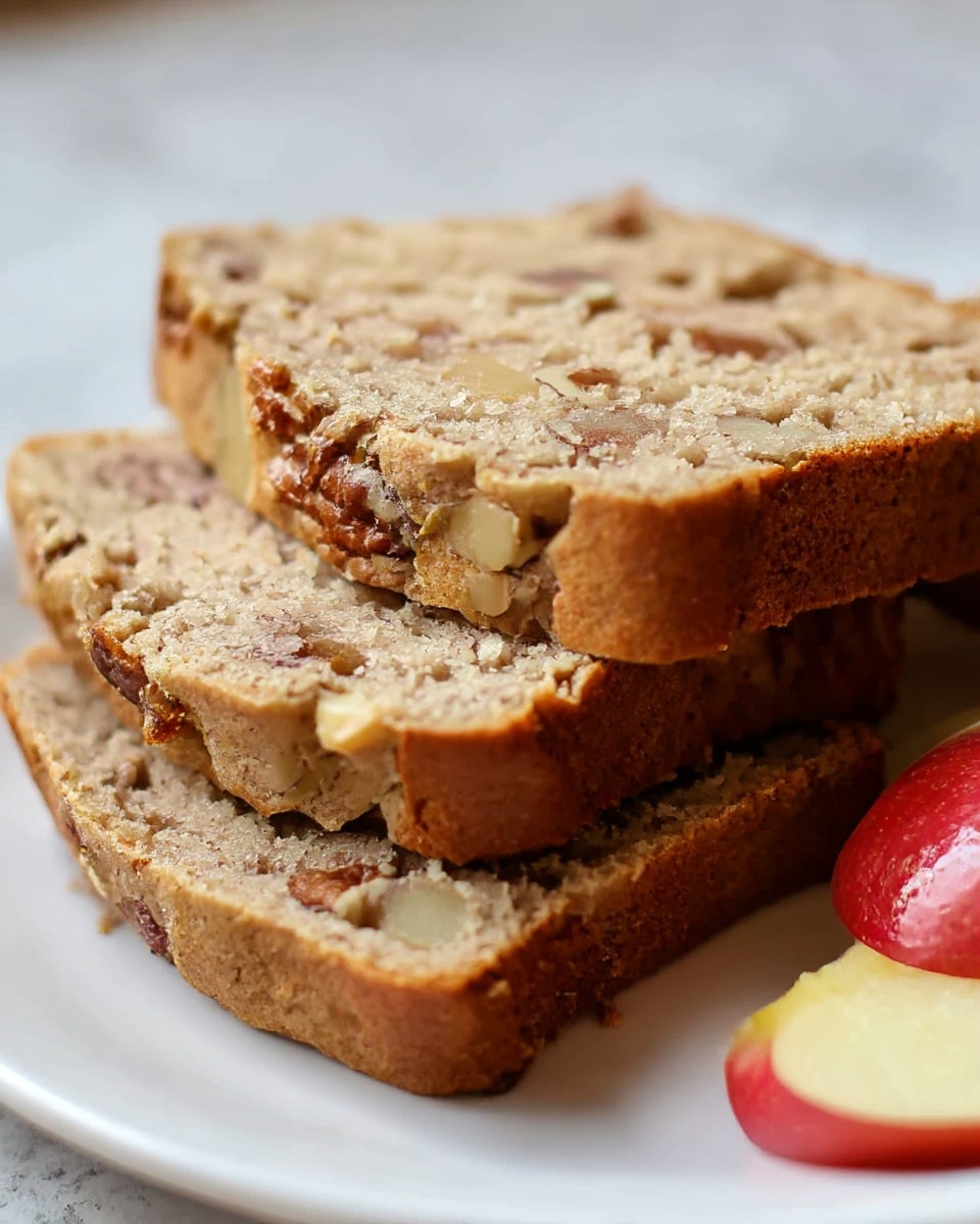 Three thick slices of nut-studded banana bread are placed side by side on a white plate, showing a dense and moist texture with visible pieces of chopped nuts throughout. The top crust is slightly crumbly and light brown, contrasting with the softer, beige inside. At the front, a slice of red apple adds a fresh pop of color, resting on the white plate against the bread. The background features a clean white marbled surface. photo taken with an iphone --ar 4:5 --v 7
