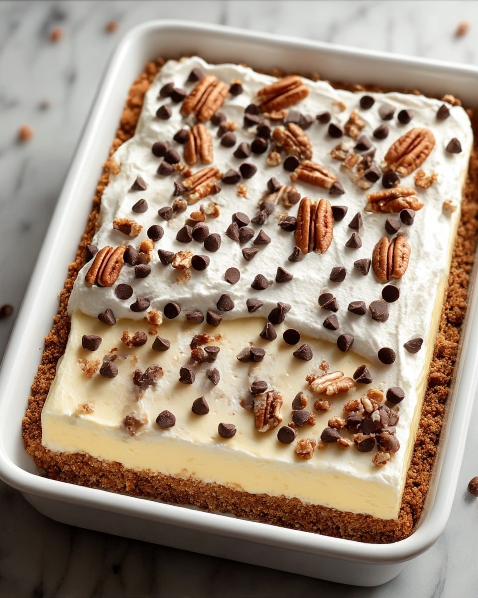 A rectangular dessert with three clear layers sits in a white baking dish. The bottom layer is a crumbly, brown crust with a rough texture. Above it is a smooth, creamy pale yellow filling that looks soft and thick. The top layer is white, fluffy whipped cream spread evenly, and this is garnished with scattered dark brown chocolate chips and broken pieces of light brown pecans, providing a crunchy contrast. The background is a white marbled surface. photo taken with an iphone --ar 4:5 --v 7