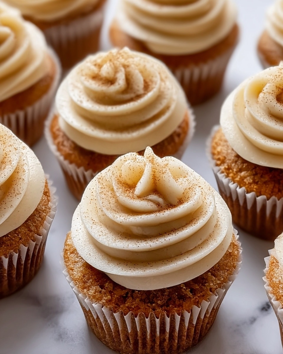 The image shows a close-up of several cupcakes on a white marbled surface. Each cupcake has two main layers: a moist brown cake base with a textured crumb and a thick, creamy white frosting on top, swirled neatly in a circular pattern. The frosting is dusted with a light sprinkle of brown cinnamon or spice powder, adding a soft contrast to the smooth cream. The cupcakes are lined with white paper liners, and the focus is sharp on the closest cupcakes, showing detailed texture on both the cake and frosting. photo taken with an iphone --ar 4:5 --v 7