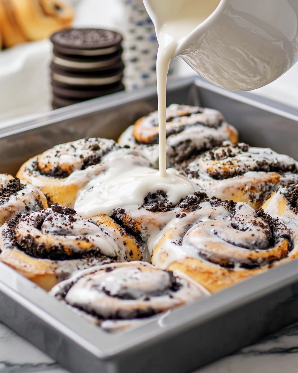 The image shows eight cookie and cream cinnamon rolls arranged closely in a square silver baking tray. Each roll has a soft golden-brown dough layer twisted with dark Oreo cookie crumbs inside, creating a strong contrast of light and dark swirls. A thick white glaze is being poured over the rolls from a small white pitcher, coating the tops with smooth, shiny icing. Crushed Oreo bits are sprinkled over the glaze, adding a rough texture. The background has a stack of Oreo cookies blurred out, and the surface is a white marbled texture. photo taken with an iphone --ar 4:5 --v 7