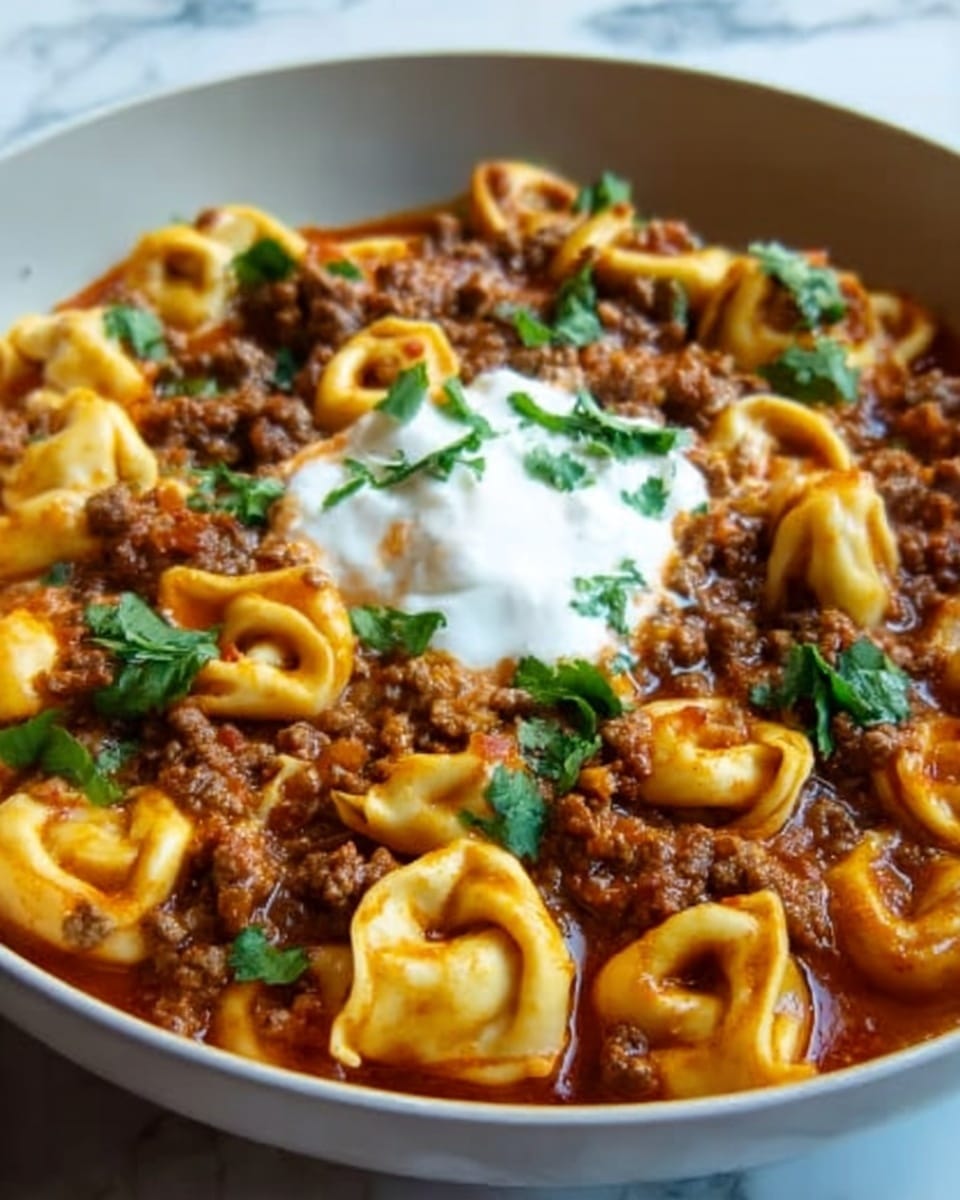 The image shows a close-up of a white bowl filled with golden yellow tortellini pasta mixed with a thick red-brown meat sauce. Small pieces of cooked ground meat are mixed throughout, giving a rich texture to the dish. On top, a dollop of white sour cream or yogurt sits in the center, surrounded by scattered bright green cilantro leaves that add fresh color contrast. The bowl is placed on a white marbled surface, highlighting the warm colors of the food. The lighting and focus capture the steam and texture clearly, making the dish look fresh and ready to eat. Photo taken with an iphone --ar 4:5 --v 7