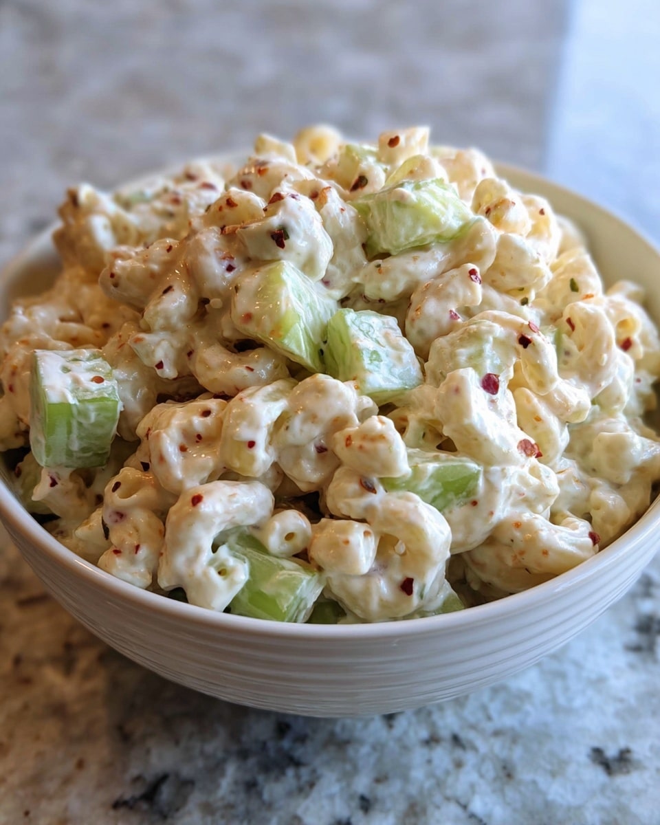 A close-up view of a bowl filled with creamy macaroni salad, showing three main layers: small pasta pieces coated in a thick white dressing, chopped celery chunks with a fresh light green color mixed evenly throughout, and dark red pepper flakes sprinkled on top adding specks of color and texture. The bowl is white and sits on a white marbled surface. The photo is taken from an angle that highlights the creamy texture and mix of ingredients with a blurred light background. photo taken with an iphone --ar 4:5 --v 7