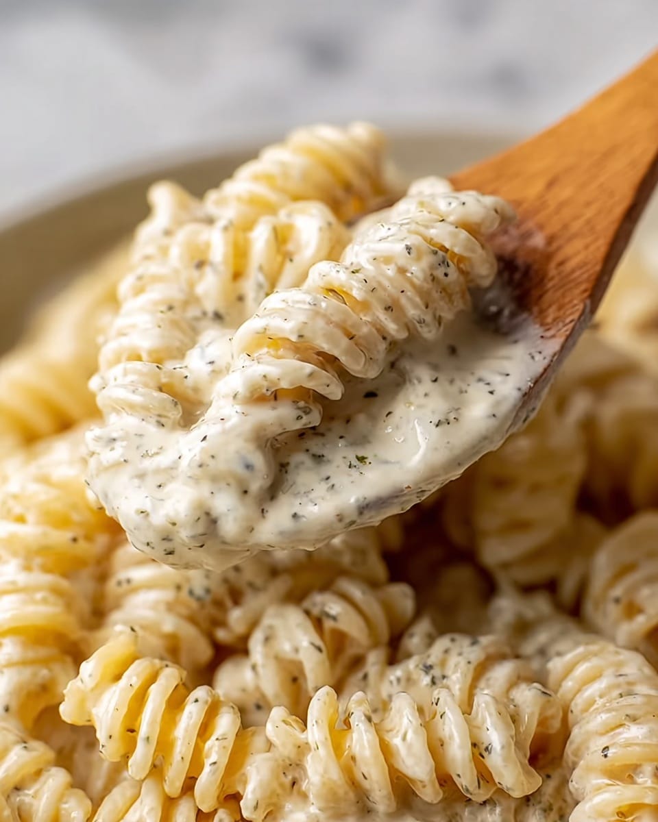 A close-up view of creamy pasta showing a wooden spoon lifting a portion of short spiral pasta fully covered in a thick, rich white sauce with small black and green herb specks evenly spread throughout. The sauce has a smooth, slightly glossy texture, coating every crevice of the spiraled pasta. The background shows more pasta in the same sauce piled together, all sitting on a white marbled surface. photo taken with an iphone --ar 4:5 --v 7