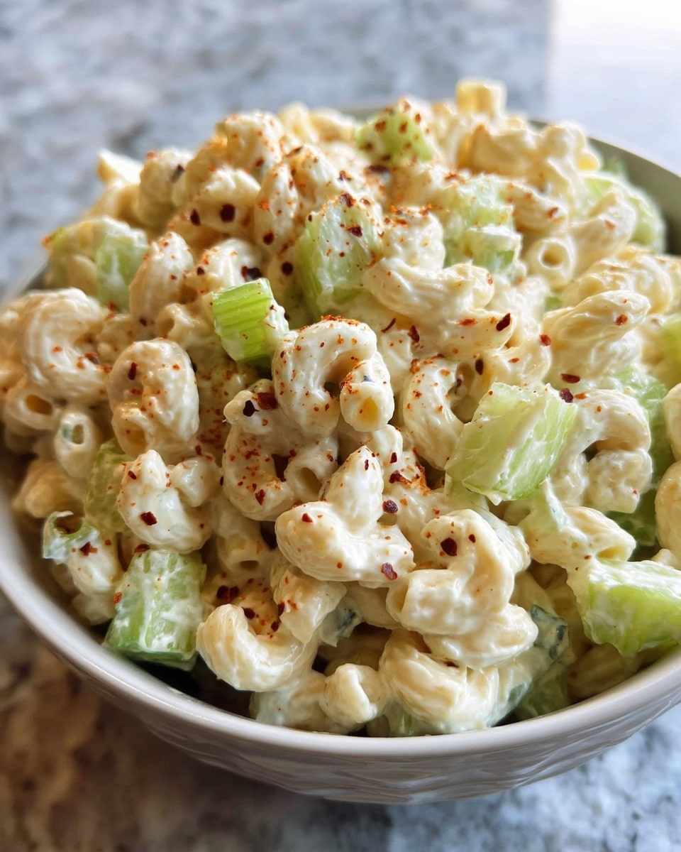 A close-up view of a white bowl filled with creamy macaroni salad, showing about two layers of elbow macaroni coated in a thick, pale white dressing with small black pepper specks scattered throughout. Mixed in are chunks of fresh, light green celery, cut into small, rough cubes, adding a crisp texture and color contrast. The salad looks rich and well-mixed, sitting on a white marbled texture surface with two blurred clear glasses in the background. photo taken with an iphone --ar 4:5 --v 7