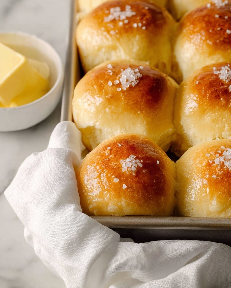 A close-up view of a baking tray filled with nine golden brown soft dinner rolls arranged in a 3x3 grid, each roll topped with coarse sea salt crystals and a shiny, buttery glaze. The rolls have a fluffy texture with slightly rounded tops and lightly browned patches showing uneven caramelization. The tray sits on a white cloth napkin over a white marbled surface, with a small white bowl containing thick yellow butter positioned at the lower left corner of the image. Photo taken with an iphone --ar 4:5 --v 7