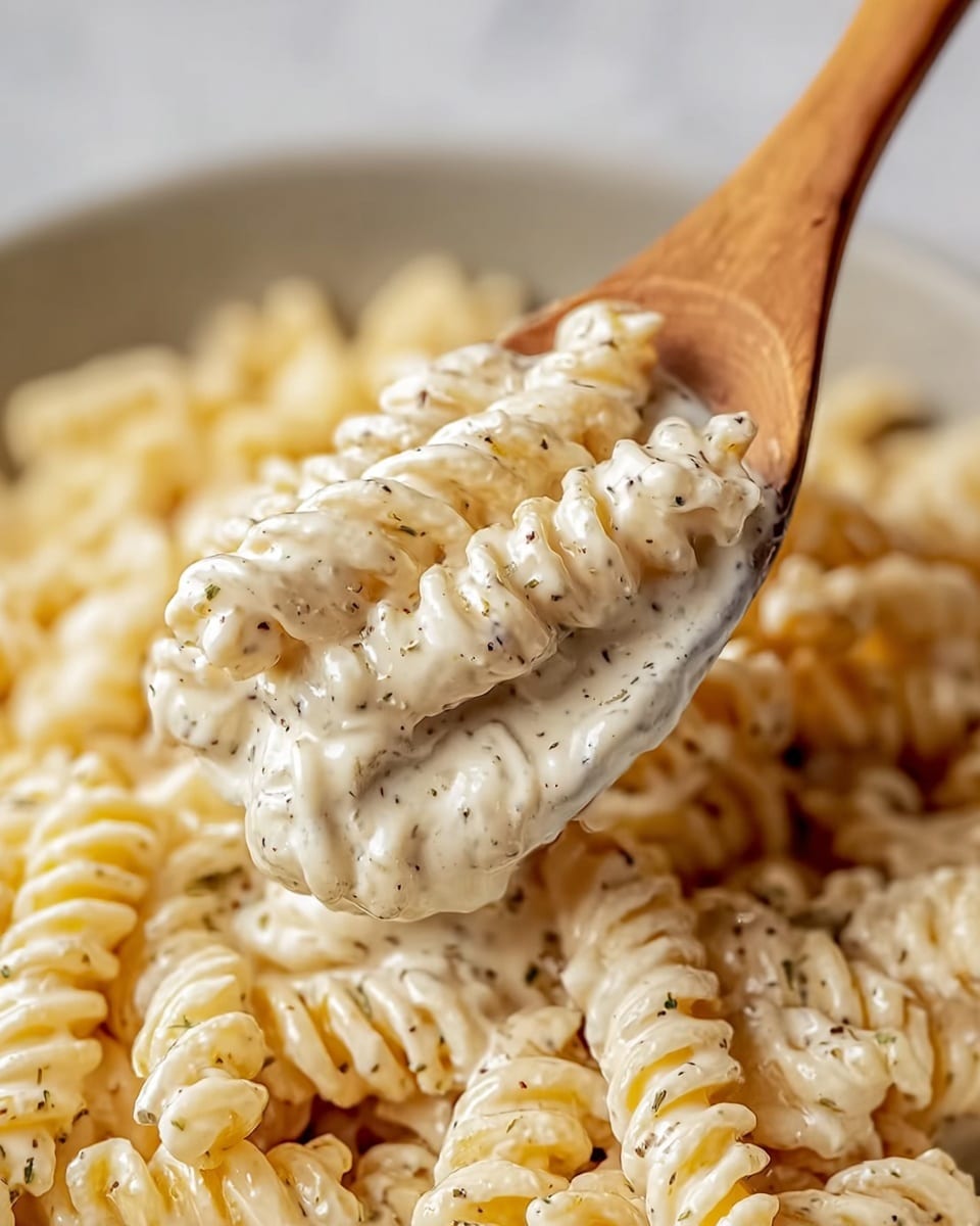 A close-up view shows a wooden spoon lifting creamy rotini pasta coated in a thick, white sauce speckled with fine black and green herbs. The pasta spirals are well covered, with the sauce clinging smoothly to each twist, showing a rich, velvety texture. The background is filled with more pasta immersed in the sauce, all resting on a white marbled surface. The scene highlights the creamy sauce’s thickness and the pasta’s soft, tender look. photo taken with an iphone --ar 4:5 --v 7