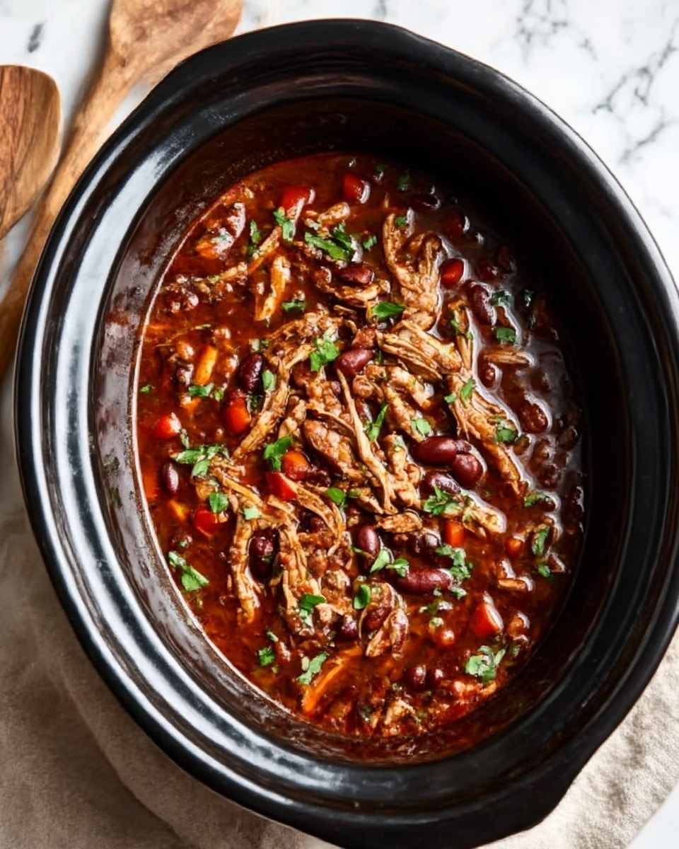 The image shows a black slow cooker filled with a rich, dark red stew. The stew contains shredded pieces of tender chicken layered on top, mixed with small chunks of red bell peppers and beans. Fresh green herbs are sprinkled over the stew, adding a touch of color contrast. The slow cooker sits on a white marbled surface with beige cloth and wooden utensils nearby. Photo taken with an iphone --ar 4:5 --v 7