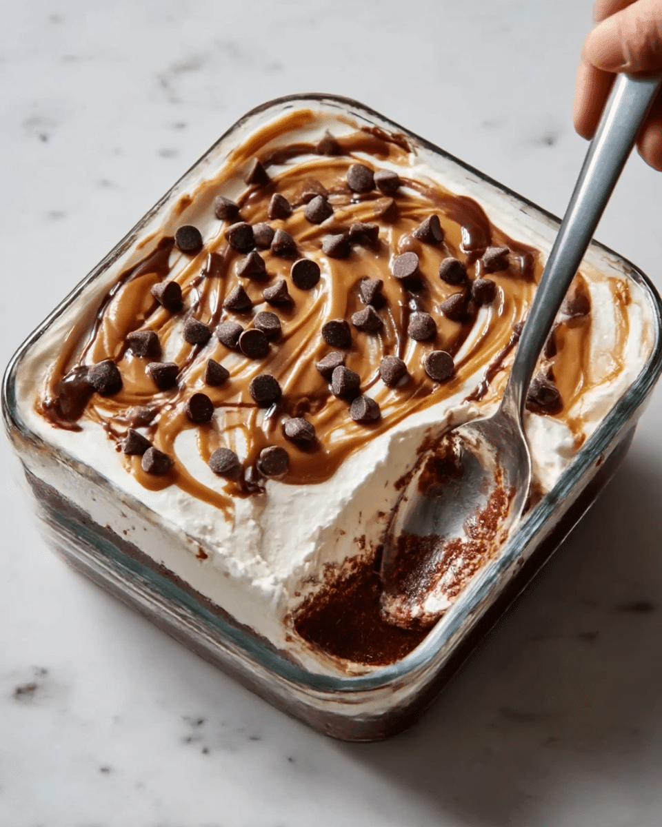 A clear glass square dish holds a layered dessert with three visible layers. The bottom layer is dark brown and smooth, likely a rich chocolate base. The middle layer is creamy white, thick and spread evenly above the dark layer. The top layer is a swirl of light brown caramel sauce spread unevenly with drizzled darker chocolate syrup creating a swirl pattern on the white cream. Scattered chocolate chips of medium size are spread across the top. A silver spoon is inserted into the right side of the dish, showing some of the dark brown layer on it. The dish rests on a white marbled surface. A woman's hand is holding the spoon. photo taken with an iphone --ar 4:5 --v 7