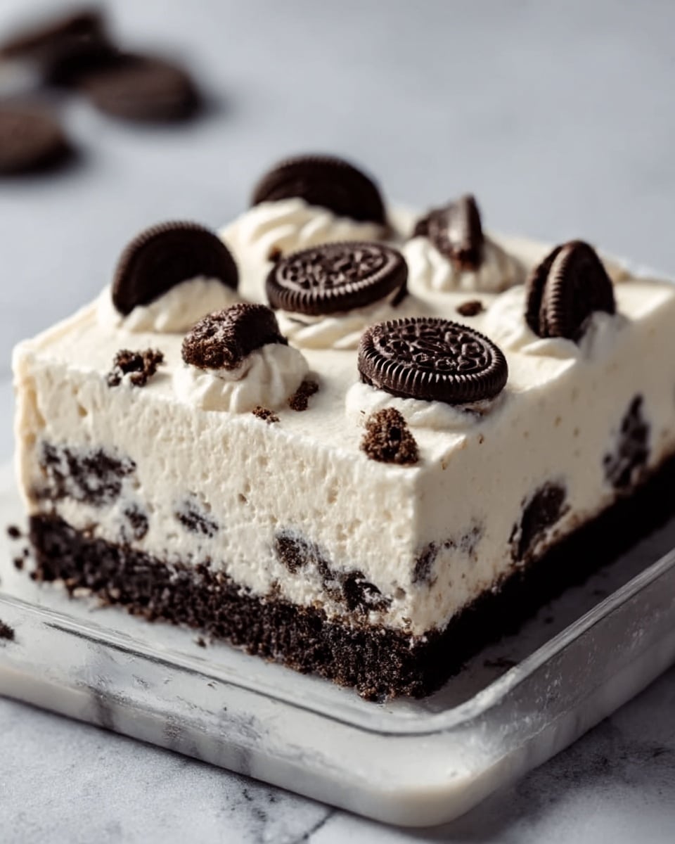 A square slice of dessert shows three distinct layers: a dark, crumbly base made of crushed cookies, a thick middle layer of smooth white cream mixed with cookie chunks, and a top layer of white cream decorated with whole round chocolate sandwich cookies evenly placed. The dessert sits on a transparent square dish on a white marbled surface. The texture looks soft and creamy with crunchy bits inside. Photo taken with an iphone --ar 4:5 --v 7