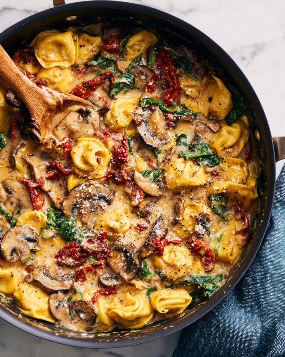 A close-up view of a black pan filled with creamy tortellini pasta in a rich sauce. The dish has three clear layers: the bottom layer is yellow tortellini with a soft, smooth texture, scattered throughout with red sun-dried tomatoes and green spinach leaves adding color and freshness. The middle layer has browned, sliced mushrooms spread around the pan edges, adding a chewy texture. The top layer is a creamy, light brown sauce speckled with black pepper and grated cheese melting softly on top. A wooden spoon is partially immersed in the pan, mixing the dish. The pan sits on a white marbled surface next to a folded blue cloth. Photo taken with an iphone --ar 4:5 --v 7