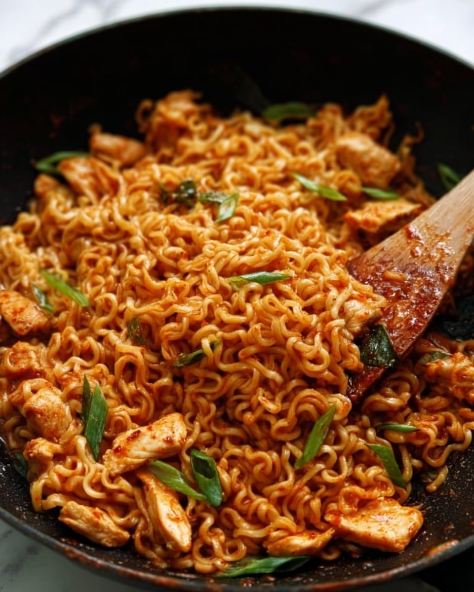 The image shows a close-up of a dark pan filled with cooked noodles mixed with pieces of chicken. The noodles are golden-brown and slightly curly, looking soft and coated with a shiny sauce. Scattered on top are small green herb leaves, adding a fresh touch of green color. A wooden spatula is stirring the noodles, held by a woman's hand on the side. The background is a white marbled texture. photo taken with an iphone --ar 4:5 --v 7