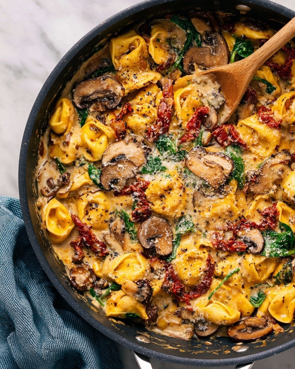 A close-up of a black pan filled with creamy tortellini pasta showing three main layers: at the base, there are soft yellow tortellini pieces; mixed throughout are sliced brown mushrooms and red sun-dried tomatoes; green spinach leaves add a fresh touch on top; the creamy sauce covers everything with a rich, slightly glossy texture; grated cheese is sprinkled lightly over the dish, melting into the sauce; a wooden spoon stirs the pasta on the left side; the pan is placed on a white marbled surface with a blue-grey textured cloth nearby. photo taken with an iphone --ar 4:5 --v 7